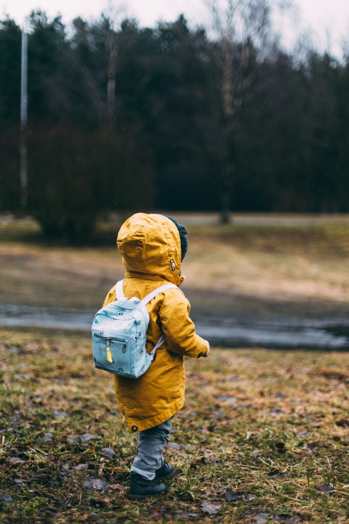 Image of a child with a yellow coat, backpack, jeans, and black shoes facing away from the camera.