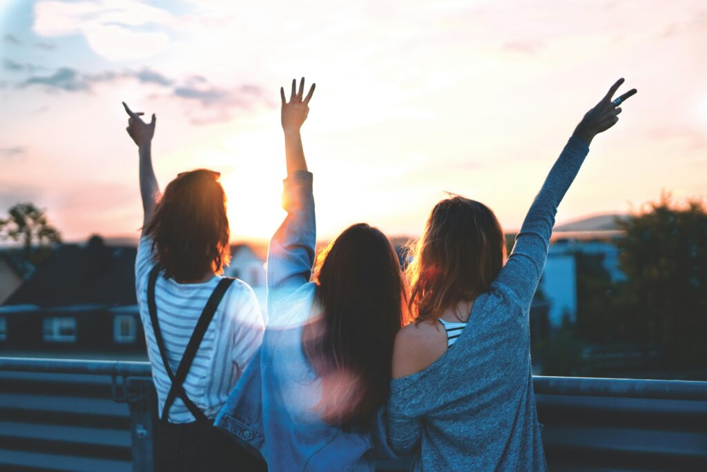 Picture of three women at sunset with their backs to the camera holding their hands in the air and embracing.