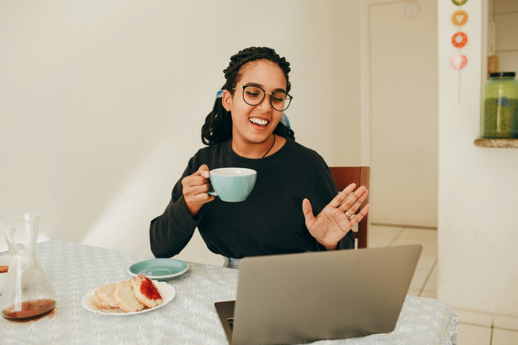 Image of a woman holding a mug and smiling and waving at her computer screen from her house.