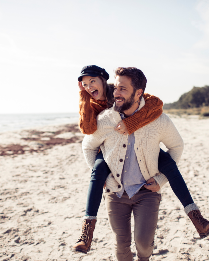 Image of a couple on the beach with the man carrying the woman on his back and both of them smiling.