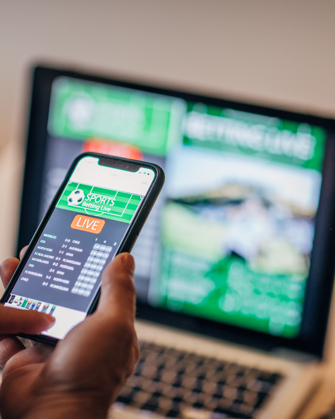 Image of a hand holding a phone with a sports betting app open and a computer in the background