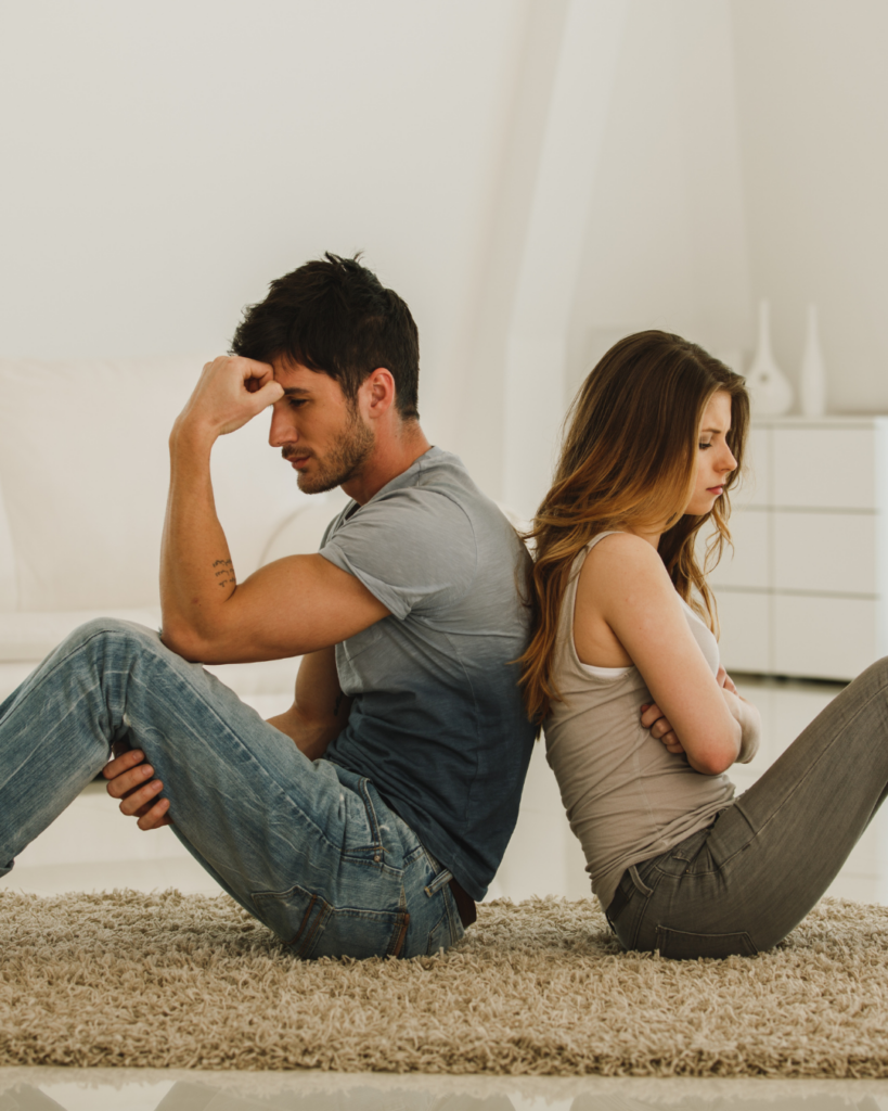 Photo of a couple sitting back-to-back on the ground and looking upset.